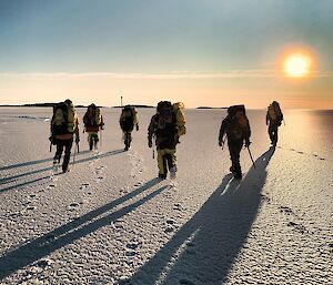 A group of six expeditioners walking away from camera over the sea ice, casting long shadows back towards the photographer.  A bright sun in the sky.