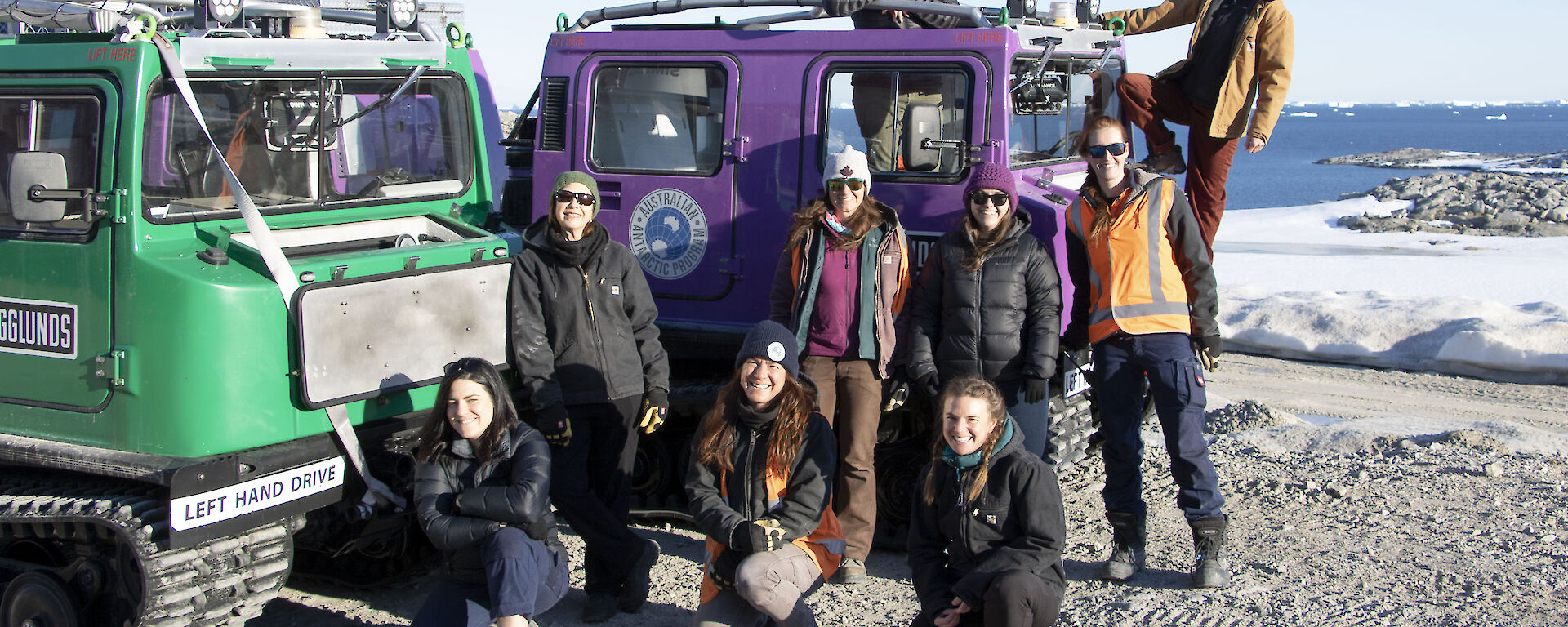 Nine women standing beside and on a purple Hagglund smiling to camera.  Snow and ice in the background