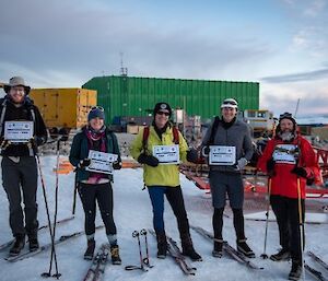 5 expeditioners stand in a line facing camera, holding up their race bibs.  Skis and poles lie beside them in the snow.