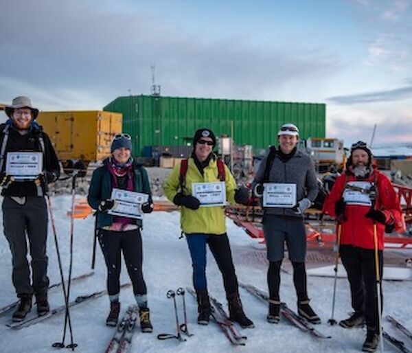 5 expeditioners stand in a line facing camera, holding up their race bibs. Skis and poles lie beside them in the snow.