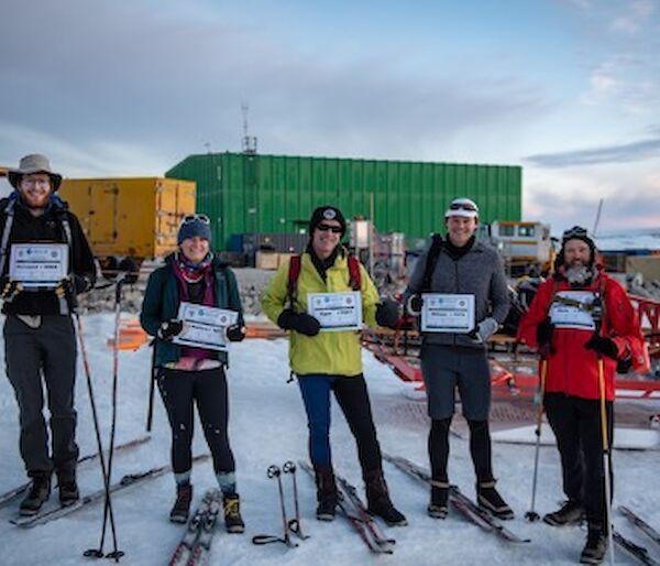 5 expeditioners stand in a line facing camera, holding up their race bibs.  Skis and poles lie beside them in the snow.