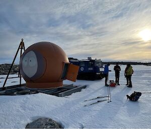 A small group of expeditioners stand outside a round field hut on the Mitchell peninsula.  The hut has been painted to look like the face of Kenny from Southpark.