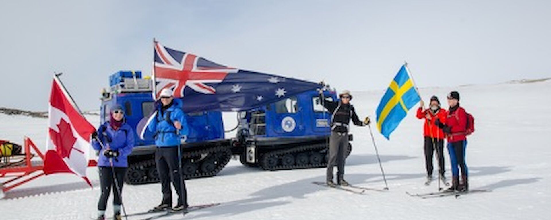 Five expeditioners on skis holding up the Australian, Canadian and Swedish flag in front of a blue Hagglund.