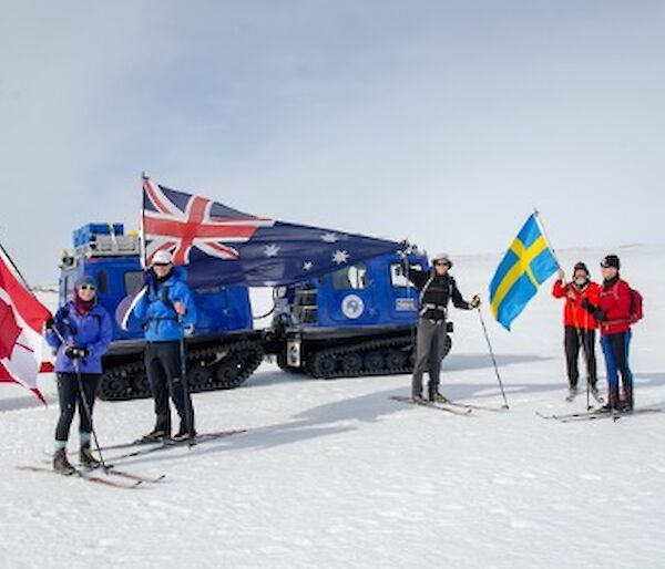Five expeditioners on skis holding up the Australian, Canadian and Swedish flag in front of a blue Hagglund.