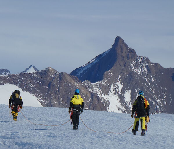 Three expeditioners wearing hard hats walking away from camera across the ice.  They are roped together for safety as they head towards large mountain range is in the distance in front of them.