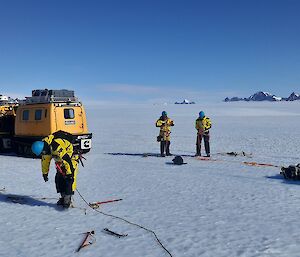 A small group of men in ice safety gear, in an icy plateau, roping themselves to each other.  Two Hagglunds behind them.