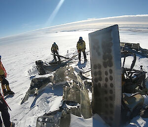 Expeditioners stand around the half buried wreckage of a Lisunov aircraft which is partially broken up in the snow.  One expeditioner is taking a photo.