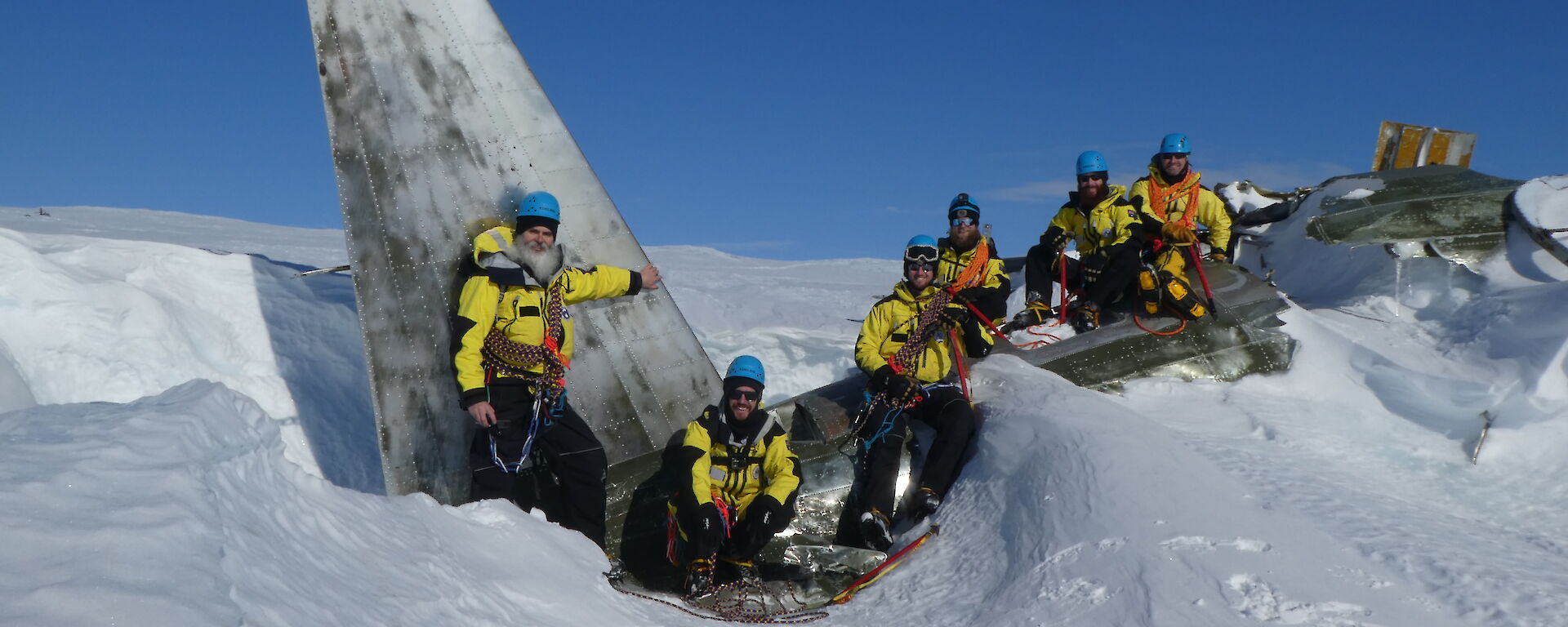 Six expeditioners sitting on the wreckage of a plane, which is half buried in snow