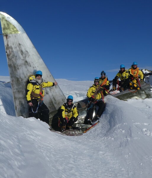 Six expeditioners sitting on the wreckage of a plane, which is half buried in snow