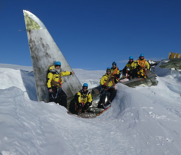 Six expeditioners sitting on the wreckage of a plane, which is half buried in snow