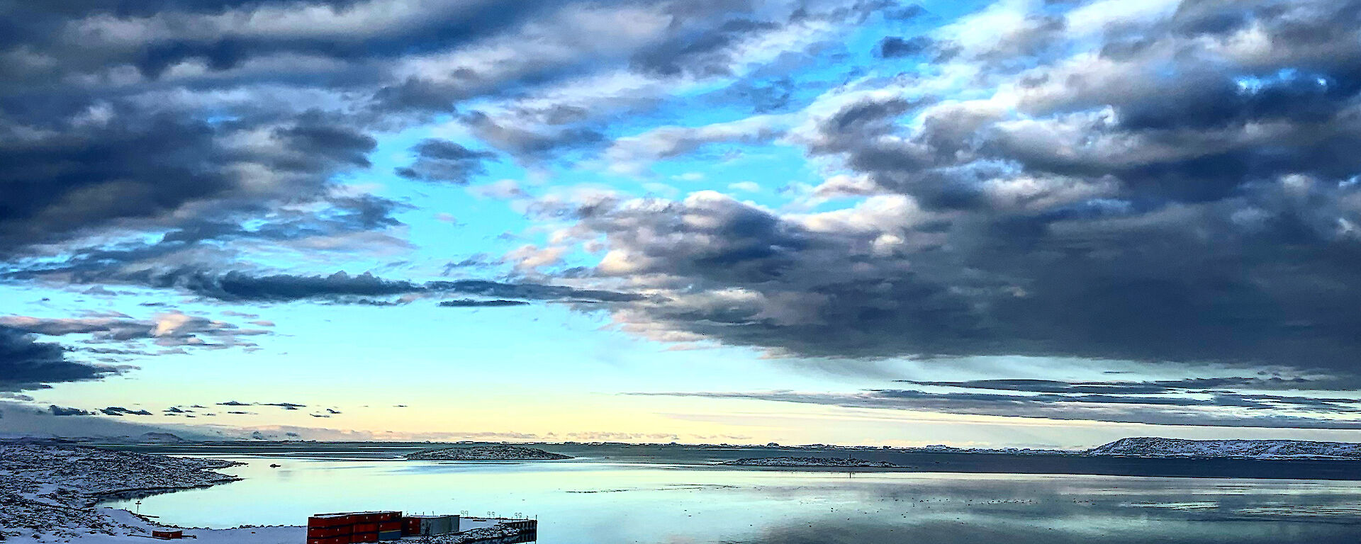 A breathtaking blue sky with gathering grey clouds reflected in the mirror smooth water below.  Snow on the shore in the foreground.