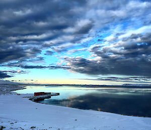 A breathtaking blue sky with gathering grey clouds reflected in the mirror smooth water below.  Snow on the shore in the foreground.