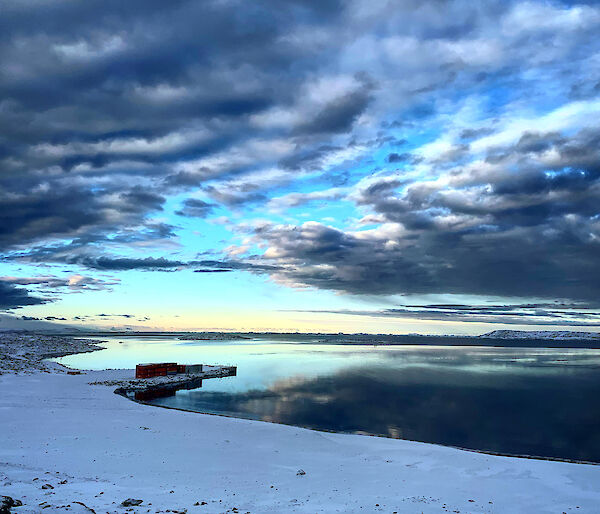 A breathtaking blue sky with gathering grey clouds reflected in the mirror smooth water below. Snow on the shore in the foreground.