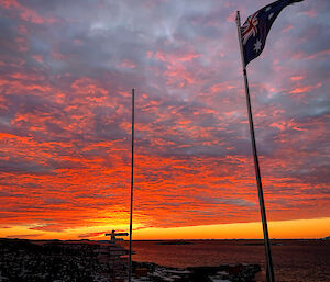 Vibrant orange sunset sky. The Australian flag atop the flagpole and Davis fingerpost sign are silhouetted against the sky