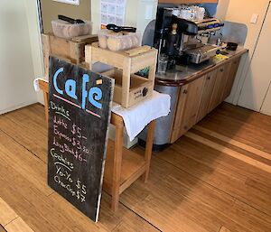 A blackboard with the words Cafe and some drink prices leans up against some wooden trolleys with biscuits on top.  A coffee machine is on a unit at th eback.
