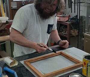 A smiling man with a bushy beard smiles to camera as he applies a coat of varnish to his hand made photo frame, in a workshop