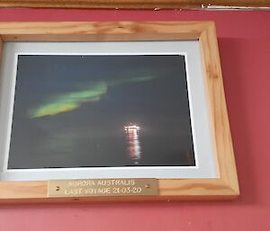 A red wall with a wooden framed photograph in the middle showing a green Aurora Australis in the sky and the lights of a boat on the water