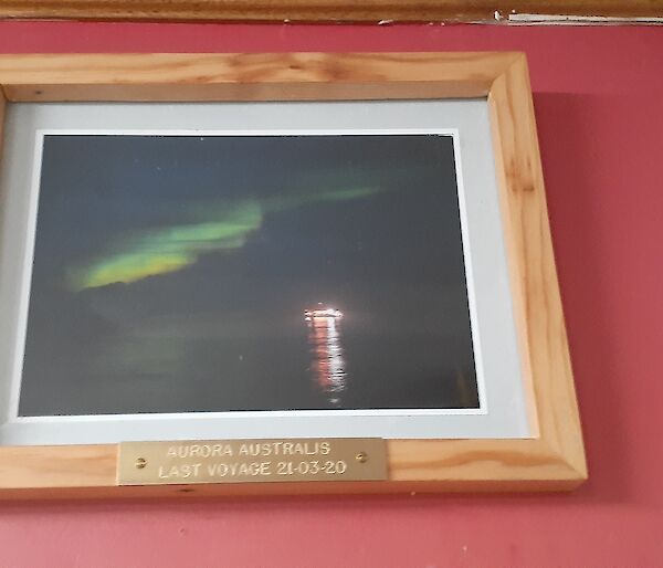 A red wall with a wooden framed photograph in the middle showing a green Aurora Australis in the sky and the lights of a boat on the water