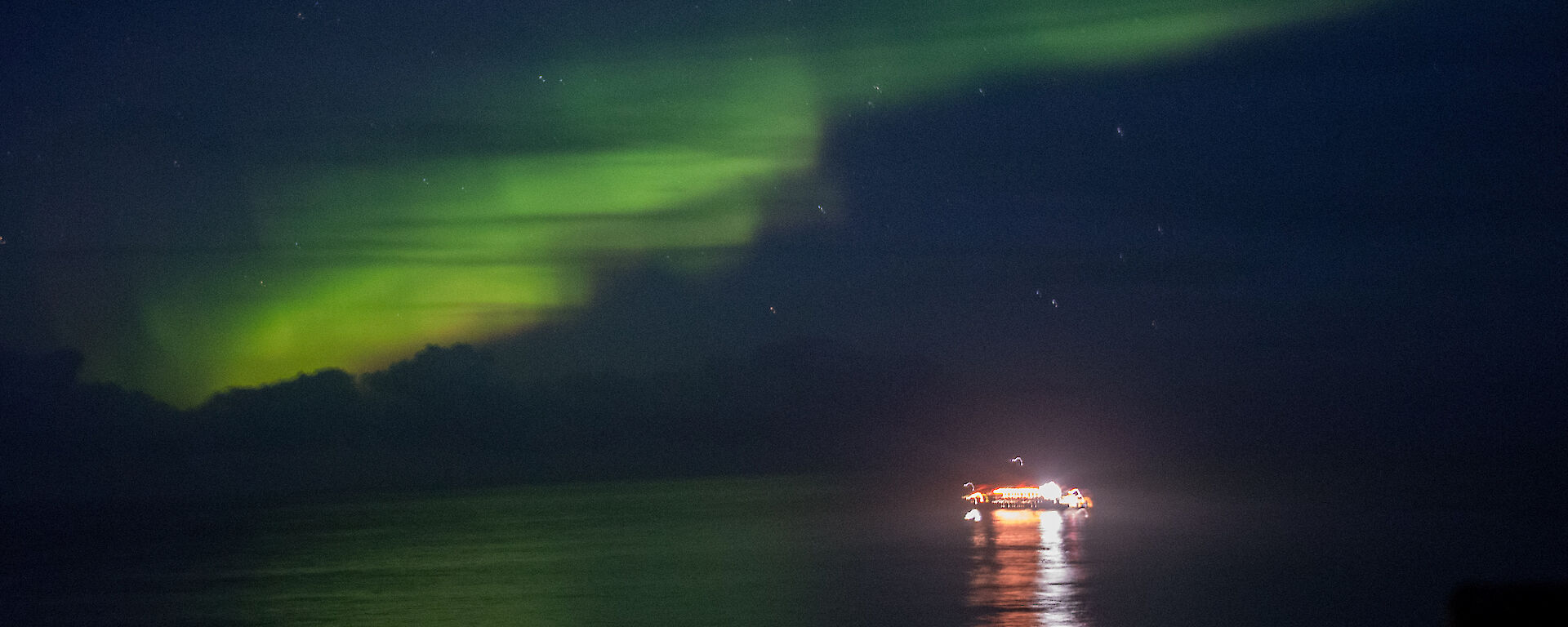 A bright green Aurora lights the dark sky.  The lights of a boat on the water are reflected in the water.