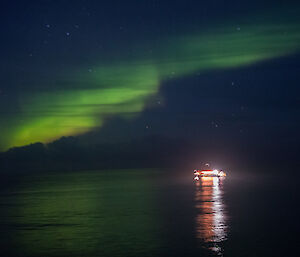 A bright green Aurora lights the dark sky.  The lights of a boat on the water are reflected in the water.