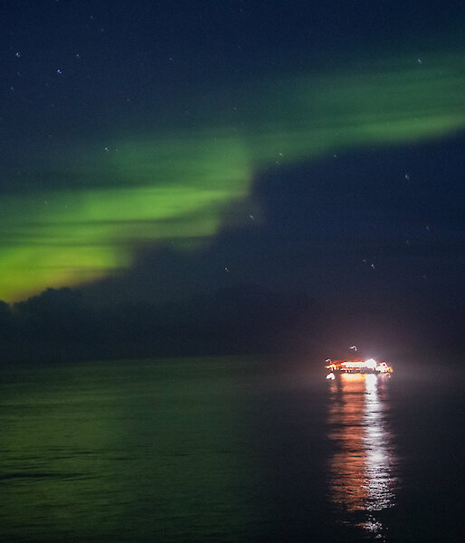 A bright green Aurora lights the dark sky.  The lights of a boat on the water are reflected in the water.