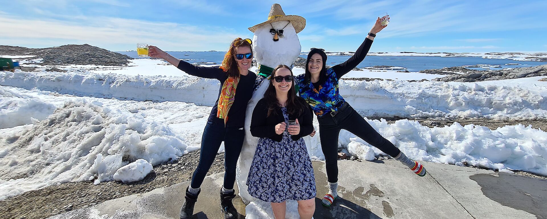 Three women stand in front of a snowman smiling.  Two are holding their drink glasses up in the air.  the snowman has a hat and sunglasses on.