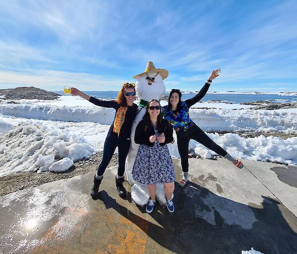 Three women stand in front of a snowman smiling.  Two are holding their drink glasses up in the air.  the snowman has a hat and sunglasses on.