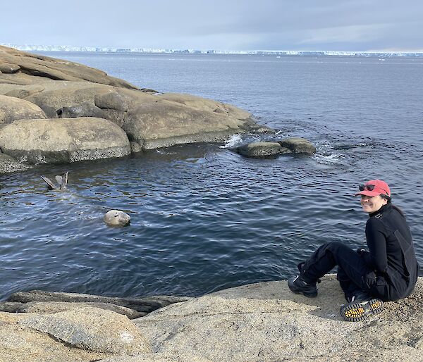 Female sitting on a rock by the shore. An seal head and tail can be seen beside her in the water. Icebergs line the horizon