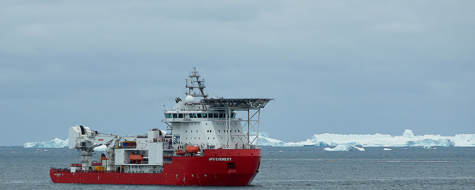 A large red and white ship sits out in a bay with icebergs on the horizon