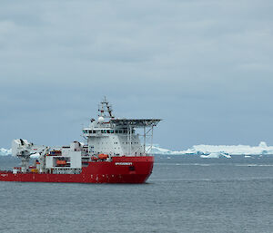 A large red and white ship sits out in a bay with icebergs on the horizon