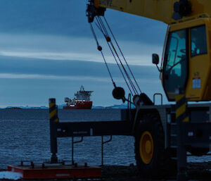 A yellow wharf crane in the foreground with a red and white ship visible behind in the bay