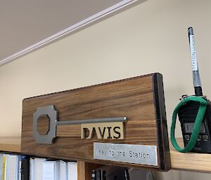 A wooden plaque with a large oversized ceremonial key with 'Davis' carved into it and a radio sitting on a bookshelf