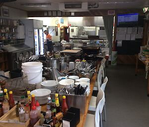 A large dining table stacked full of kitchen equipment, crockery and sauces.  A man can be seen in the kitchen in the background.