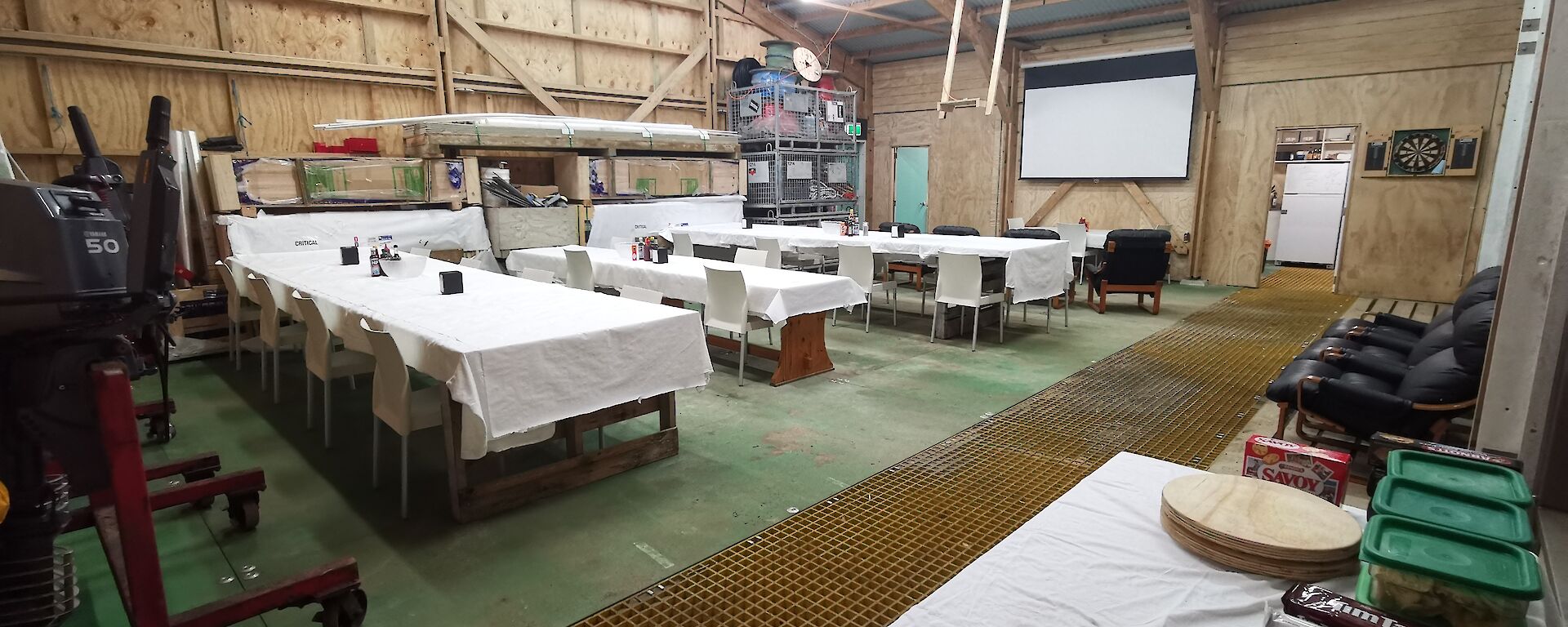 A large shed with wooden walls and a corrugated room, set up with dining tables covered in white table cloths, loungers facing a large white screen on the wall and a snack table in the  foreground