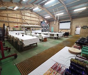 A large shed with wooden walls and a corrugated room, set up with dining tables covered in white table cloths, loungers facing a large white screen on the wall and a snack table in the  foreground