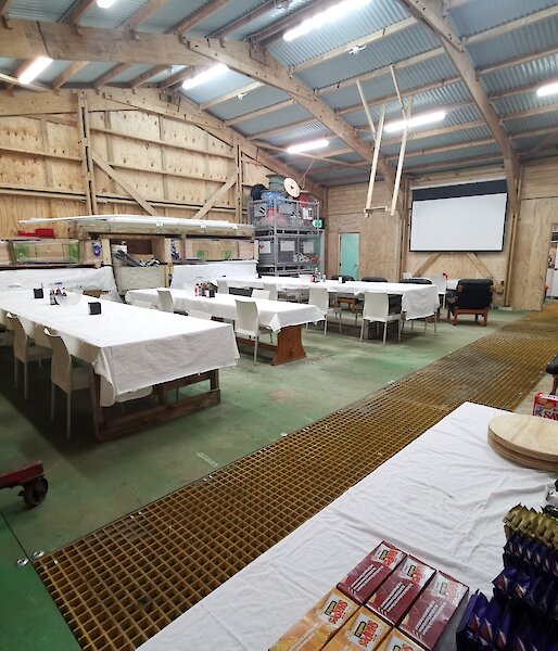 A large shed with wooden walls and a corrugated room, set up with dining tables covered in white table cloths, loungers facing a large white screen on the wall and a snack table in the  foreground