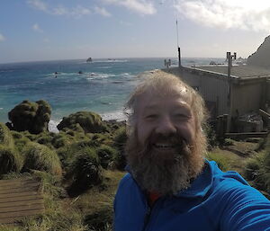 A smiling selfie of a bearded expeditioner with green tussocks and grass behind him, a shed to the right and a beautiful turquoise sea