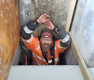 A tradesman from the waste up, with his hands above his head and a headlamp on emerges from a small enclosed space