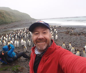 A bearded man in a baseball cap smiling in a selfie.  Behind are a large number of king penguins and another expeditioner sitting on the ground taking a photo.