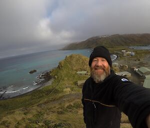 A smiling, bearded man taking a selfie from the top of a hill looking back down to the greenery of the island and the turquoise waters surrounding the island