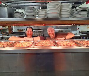 Two mean smiling behind a bain marie filled with pizzas