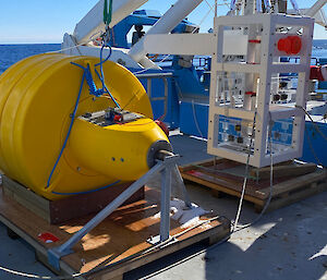 A yellow float and a white metal frame with some cameras attached, sitting on wooden pallets on a ship's deck.