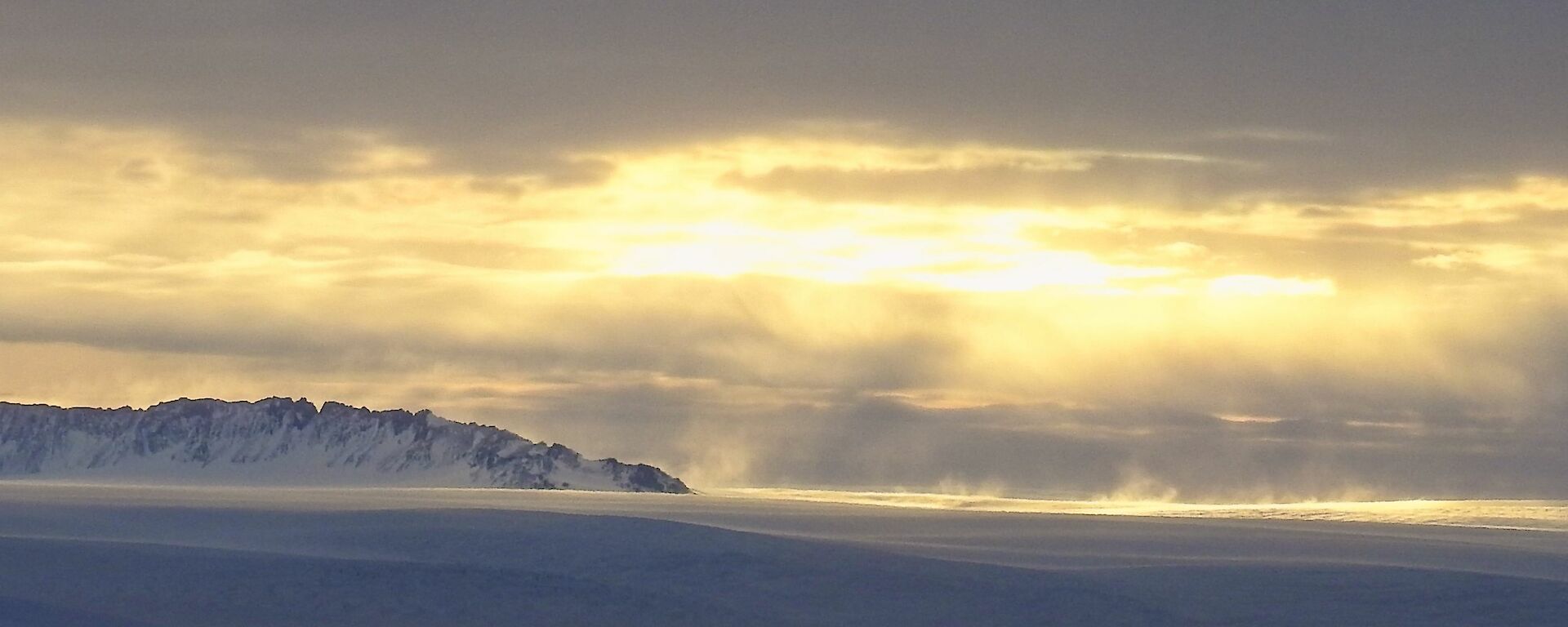 A panoramic image of  the ridge of a mountain popping out the top of the snow and ice.  Wind is blowing the surface snow across shot and the sun breaks through the clouds above.