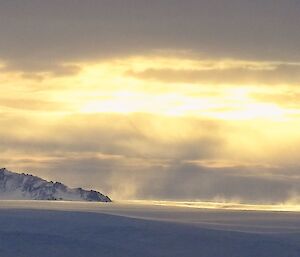 A panoramic image of  the ridge of a mountain popping out the top of the snow and ice.  Wind is blowing the surface snow across shot and the sun breaks through the clouds above.