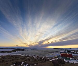 Clouds make tracks in the sky from the horizon to the foreground.  Sheds and buildings on the ground below.