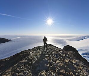 Silhouette of a man standing on a ridge with snow and clear blue sky behind.  The sun is at the centre of the image.