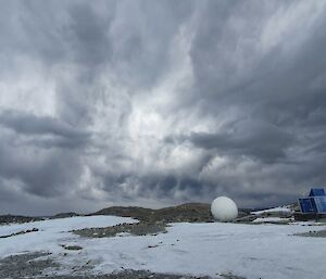 A snowy landscape with a radome and small blue shed on the horizon.  Dramatic grey storm clouds in the sky.