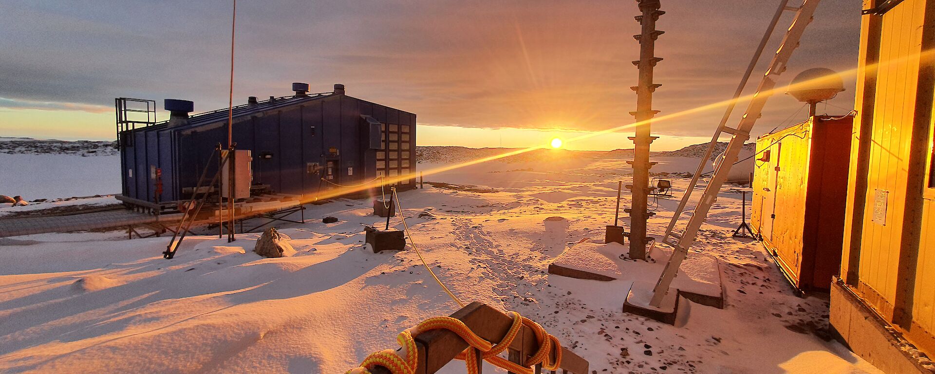 A handrail in the foreground with station sheds and equipment bathed in a yellow glow from the sun coming over the horizon.  Snow on the ground.