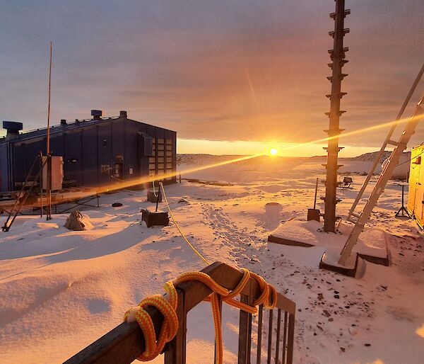 A handrail in the foreground with station sheds and equipment bathed in a yellow glow from the sun coming over the horizon.  Snow on the ground.