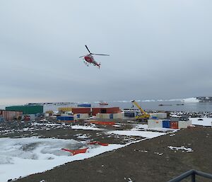 A helicopter in the sky with sheds, containers and a crane on the land below at Davis Station helipad